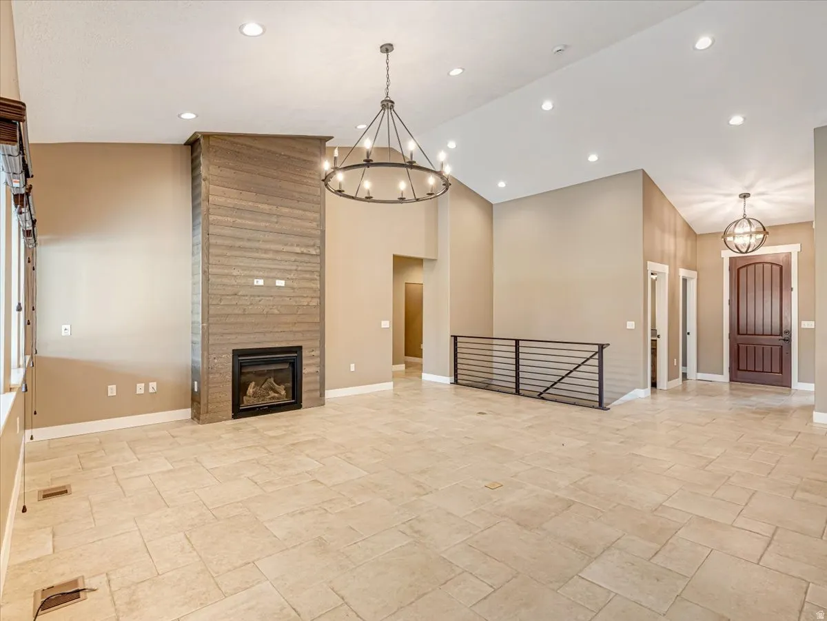 Spacious room featuring vaulted ceilings, a prominent fireplace with a wood-finish surround, large format tile flooring, a contemporary chandelier, and a metal railing