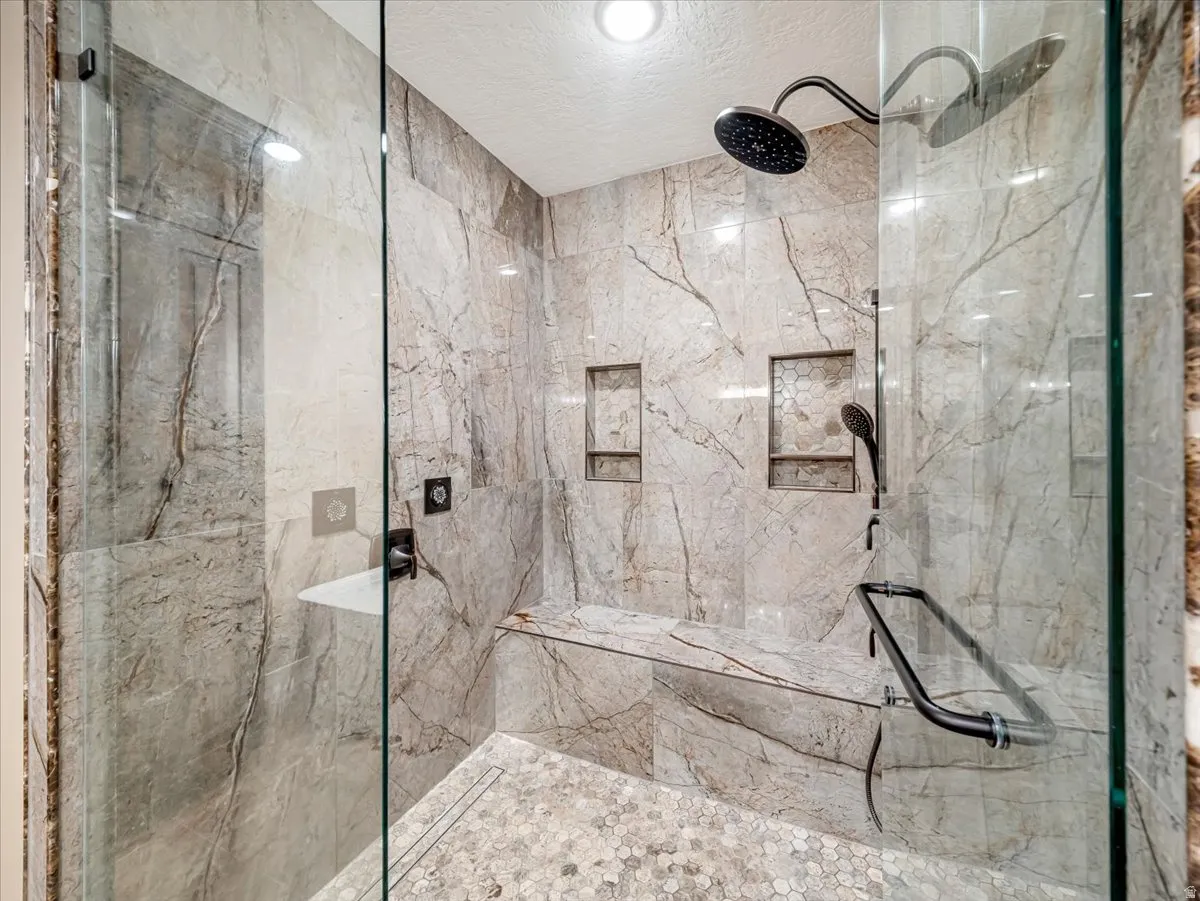 Glass-enclosed shower featuring large format veined wall tiles, a built-in bench, two shower niches, and a rainfall showerhead with an additional handheld sprayer