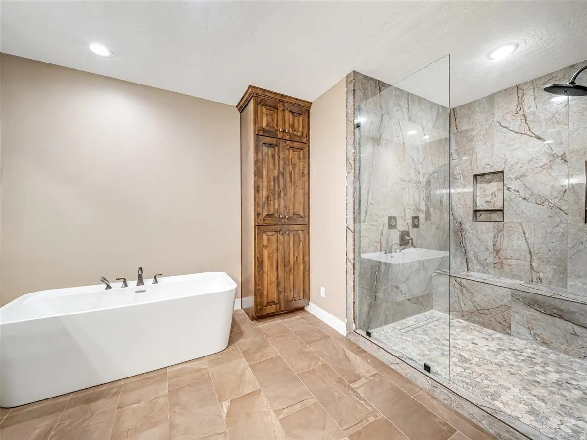 Spacious bathroom featuring a freestanding soaking tub with deck-mounted fixtures, a glass-enclosed shower with stone-look tile walls, a rain shower head, and a built-in shower bench