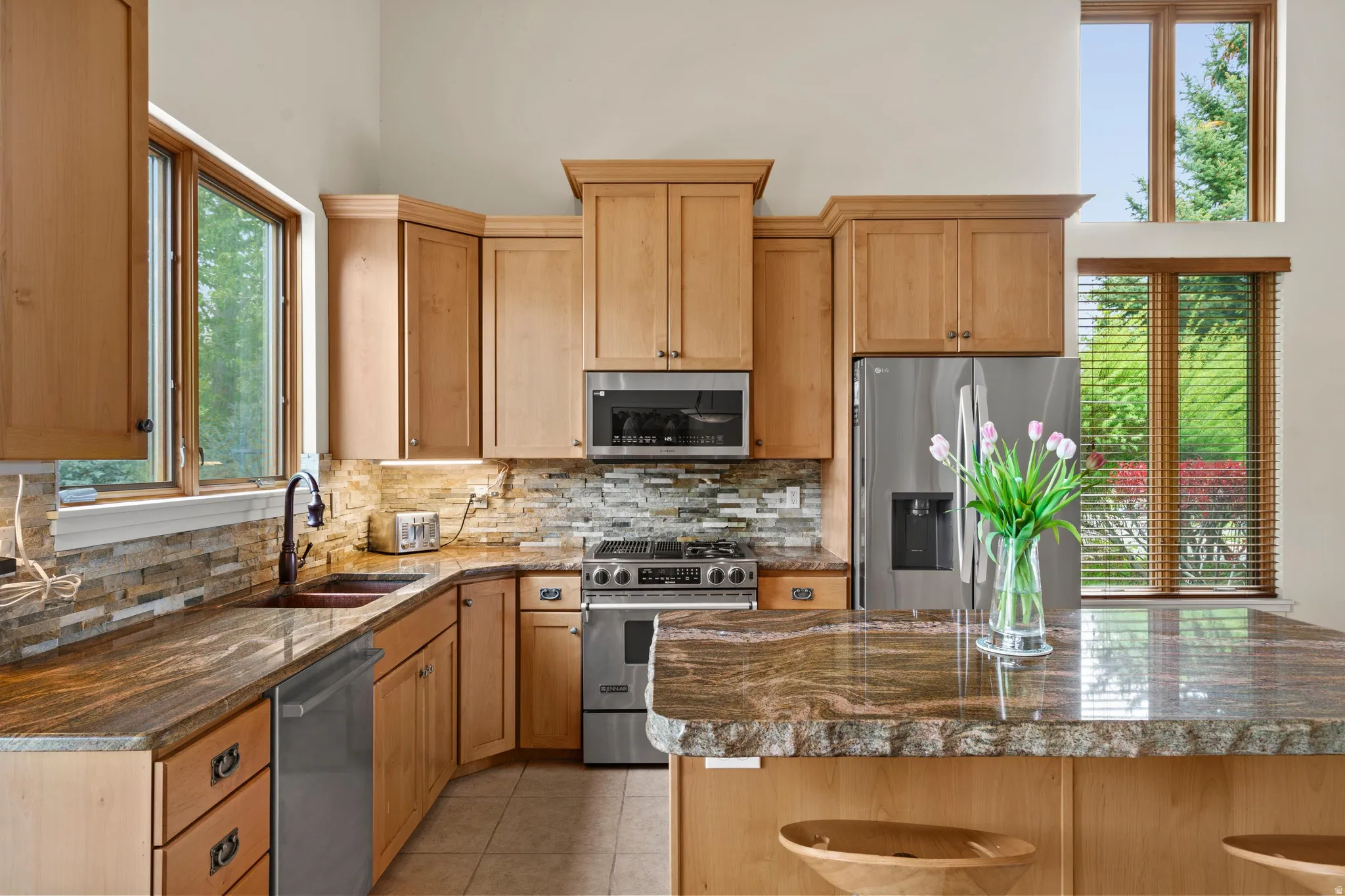 Kitchen featuring light wood cabinetry, stone slab countertops, a mosaic tile backsplash, stainless steel appliances, and a central island with bar seating