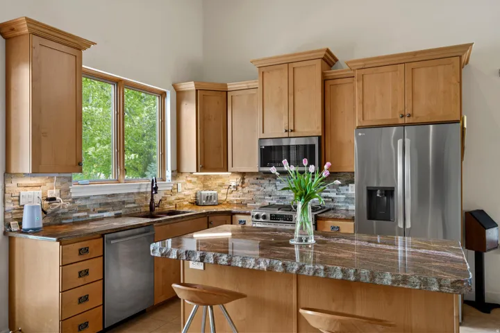 Kitchen featuring light wood cabinetry, stone countertops, stainless steel appliances, a tile backsplash, and a large window