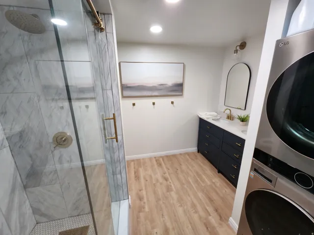 Spacious bathroom featuring a glass-enclosed shower with marble-look tile, wood-finish flooring, a long navy vanity with gold hardware, a white countertop, and a stacked washer and dryer