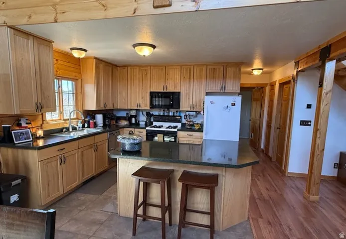 Kitchen featuring wood cabinetry, dark countertops, a central island with seating, a stainless steel sink with a gooseneck faucet, and a combination of tile and wood-finish flooring
