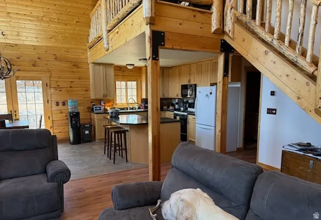 Open-concept living space featuring wood-finish walls and ceilings, a kitchen with dark countertops and light cabinetry, a built-in kitchen island with bar seating, and a log staircase leading to an upper loft