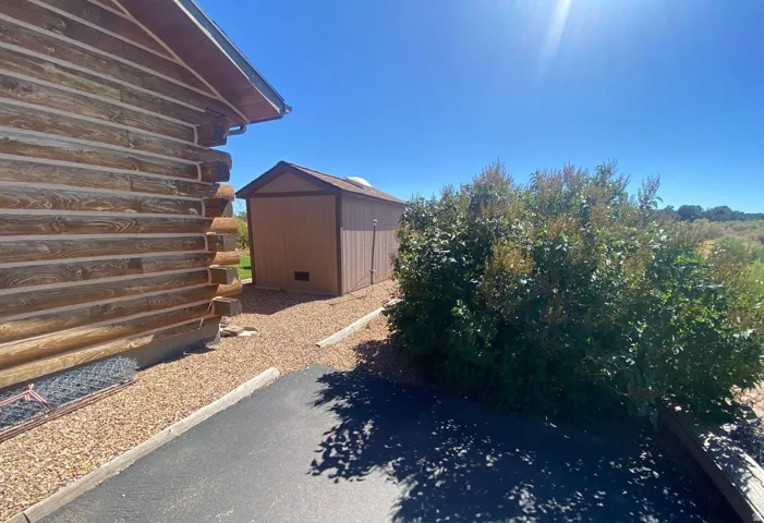 Log-style exterior featuring visible chinking, a brown storage shed, gravel landscaping, and mature shrubbery