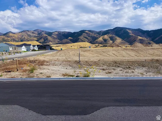 Expansive undeveloped land featuring a mountain backdrop, partially paved road, and established residential structures in the distance