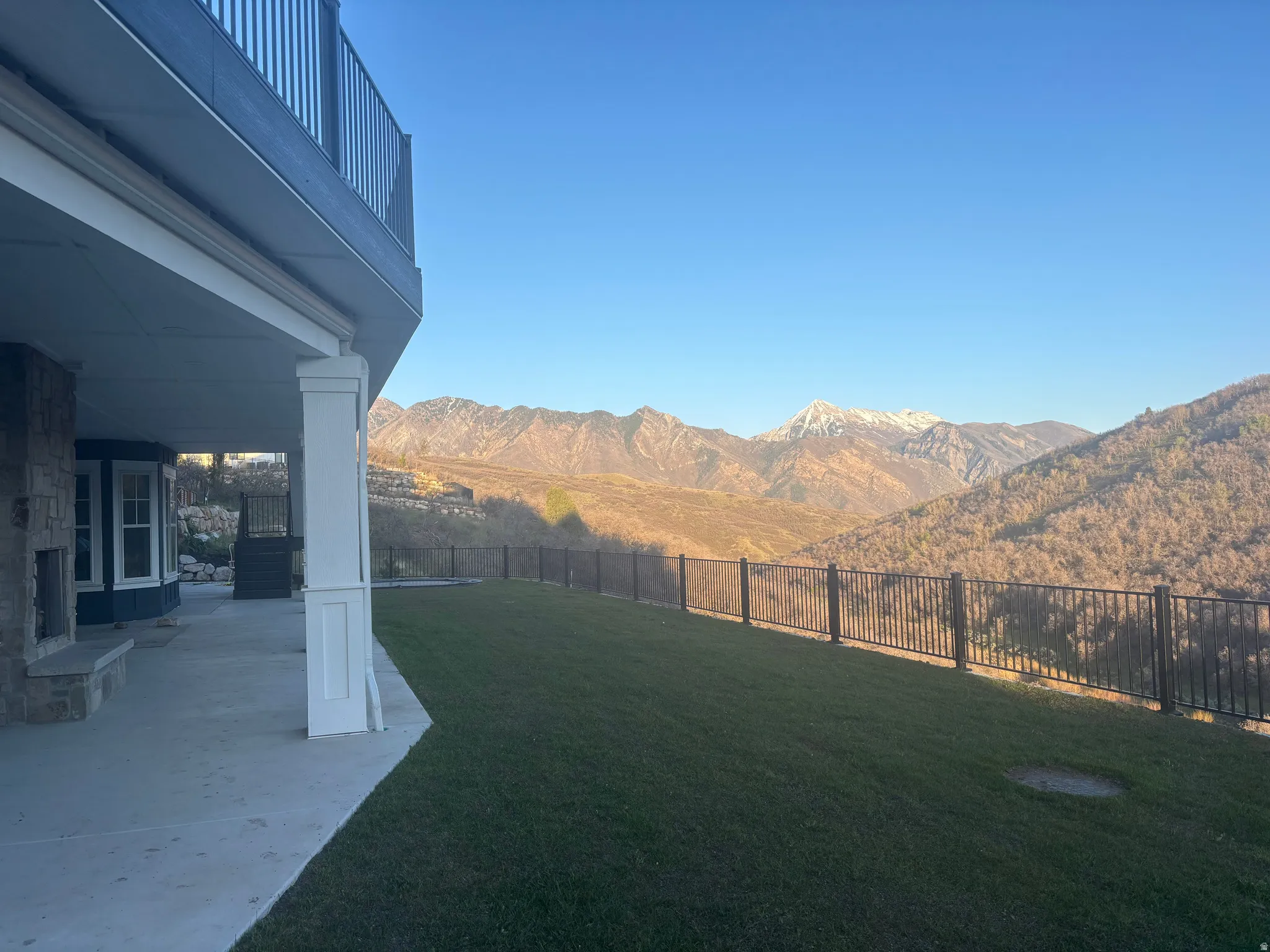 Spacious outdoor patio with stone fireplace, leading to a landscaped lawn with mountain scenery and a black metal fence