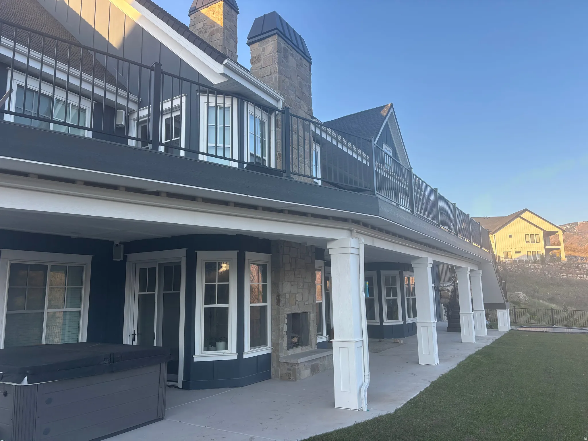 Expansive outdoor living area featuring a covered patio with a stone fireplace, an upper-level balcony with metal railings, a manicured lawn, and large windows