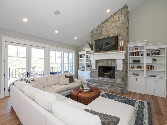 Great room featuring a stone fireplace with a substantial hearth, built-in shelving, wood-finish flooring, and double French doors opening to an exterior deck