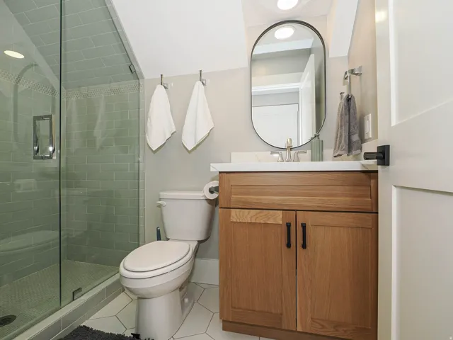 Full bathroom featuring a glass-enclosed shower with subway tile surround, a wood-finish vanity with a white countertop, an oval mirror, and hexagonal floor tiling