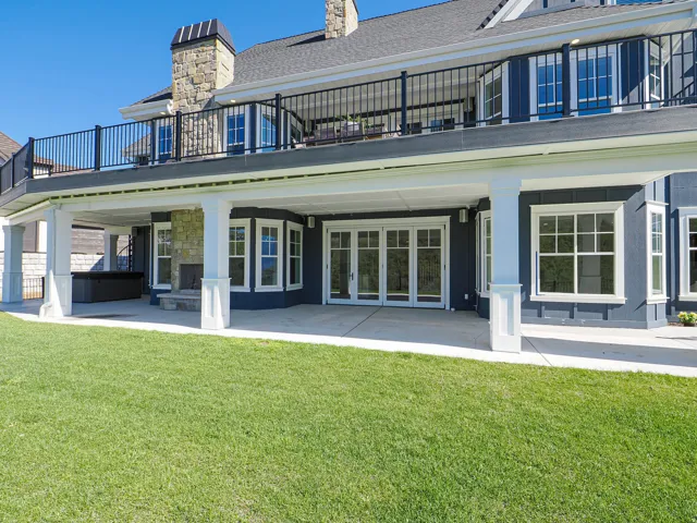 Rear exterior featuring a covered patio with stone fireplace, an upper-level balcony with black railing, and a private spa
