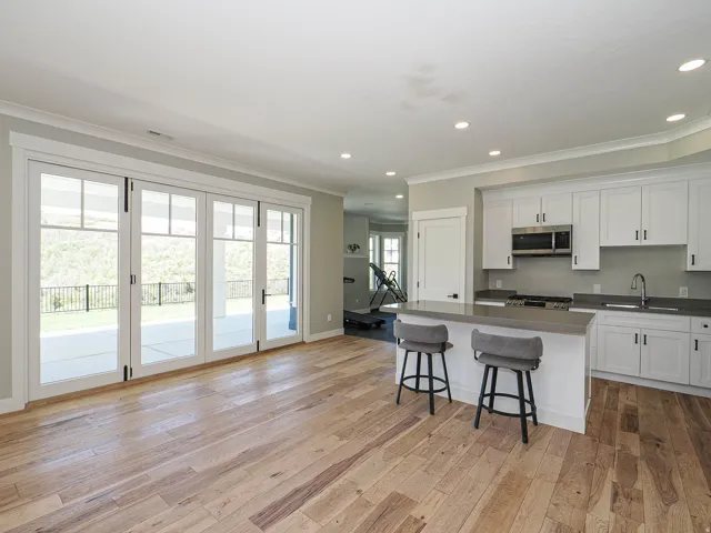 Open-concept living space featuring wood-finish flooring, a kitchen island with seating, white cabinetry, stainless steel appliances, and large glass paneled doors opening to an outdoor patio