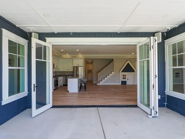 Expansive bi-fold glass doors open to a spacious interior featuring wood-finish flooring, a kitchen with white cabinetry and stainless steel appliances, and a staircase with white risers and wood treads