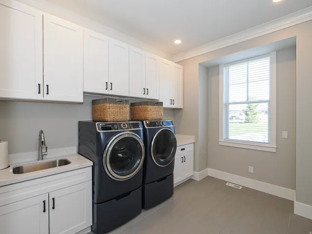 Laundry room featuring white shaker-style cabinetry with matte black hardware, a stainless steel utility sink with a pull-down faucet, light-toned countertops, and light gray tile flooring