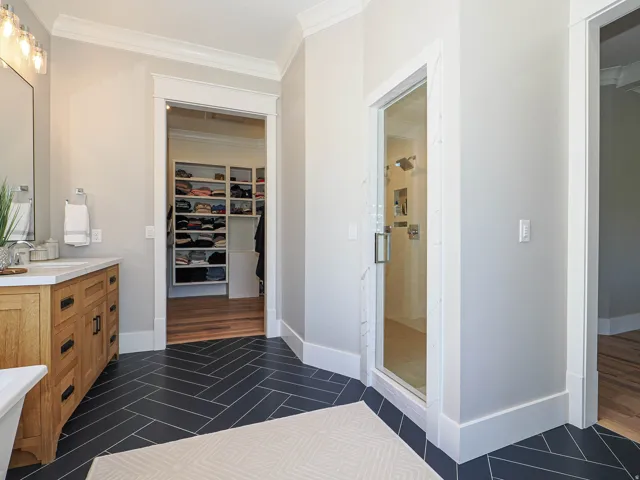 Bathroom featuring herringbone-patterned tile flooring, a glass-enclosed shower with multiple showerheads, a dual-sink vanity with a light-colored countertop, and recessed built-in shelving