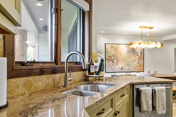 Kitchen featuring a double basin stainless steel sink with a gooseneck faucet, light brown granite countertops, diagonal-set tile backsplash, and sage green cabinetry with dark hardware
