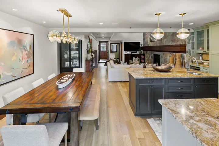Open-concept living space featuring light wood-finish flooring, a long dining table under a linear brass chandelier, a kitchen island with stone countertops, and a stone fireplace with a wood mantle