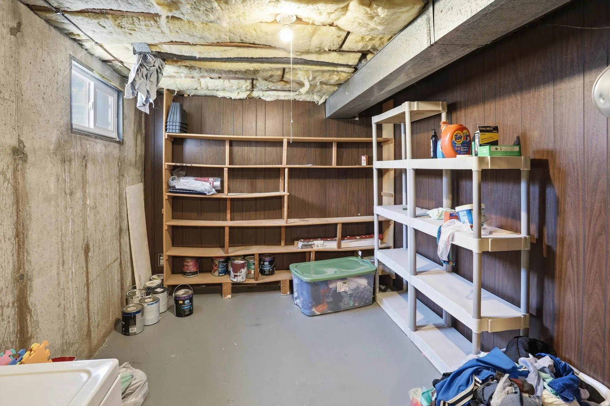 Basement area featuring a concrete wall, a small window, wood paneling, wood shelving, and a painted concrete floor