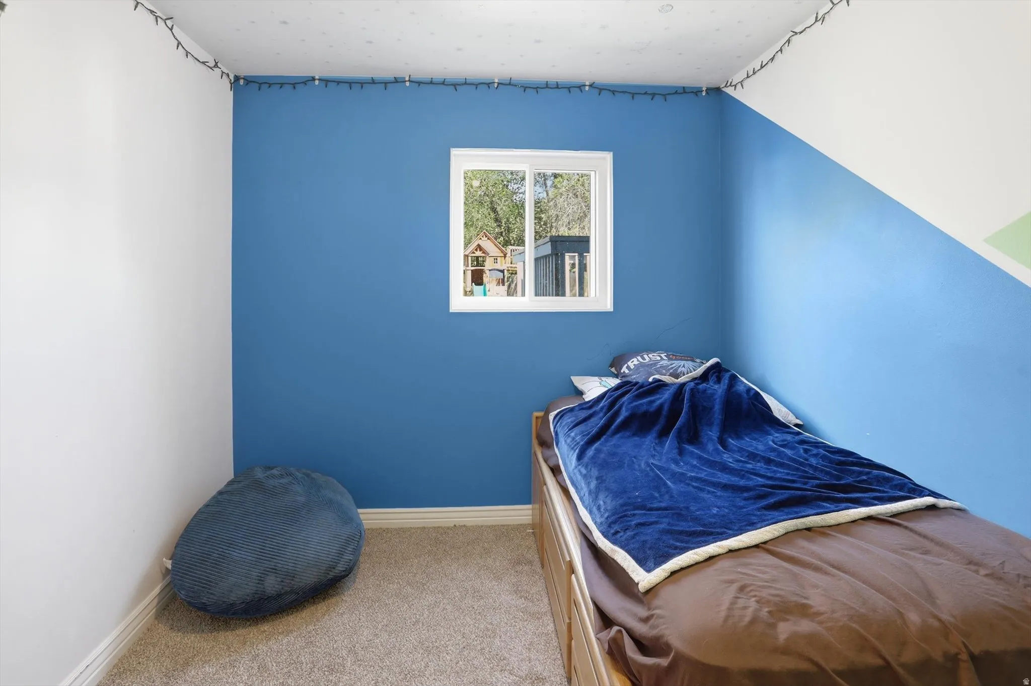 Carpeted room featuring a white-framed window, painted accent walls, white baseboards, and string lighting along the ceiling perimeter