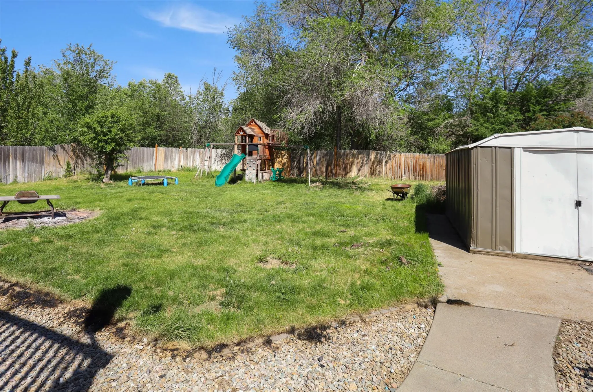 Expansive grassy yard featuring a wood privacy fence, mature trees, a paved pathway, and a detached storage shed