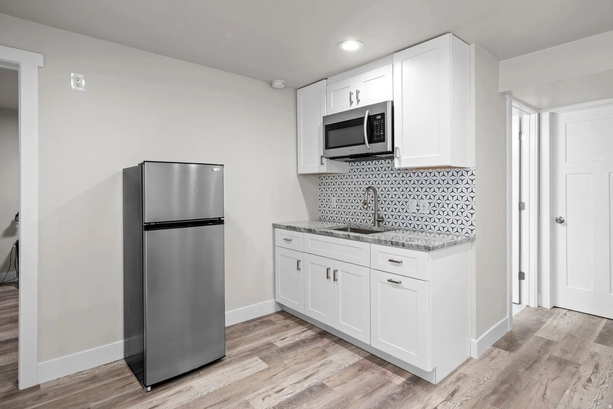 Basement kitchenette featuring white cabinets, tile backsplash, sink, microwave, and fridge.