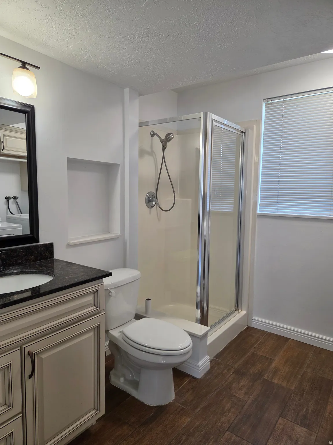 Full bathroom featuring a walk-in shower with glass enclosure, single vanity with dark granite countertop, light-colored cabinetry with dark hardware, and wood-finish flooring