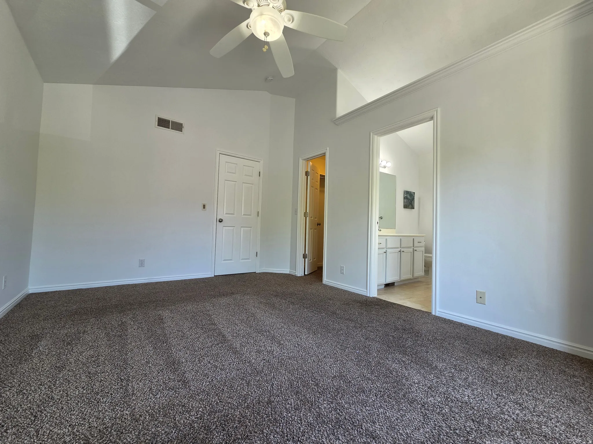Spacious room featuring vaulted ceilings, a ceiling fan, plush carpet flooring, a paneled interior door, and a dedicated entry to a bathroom with a dual vanity