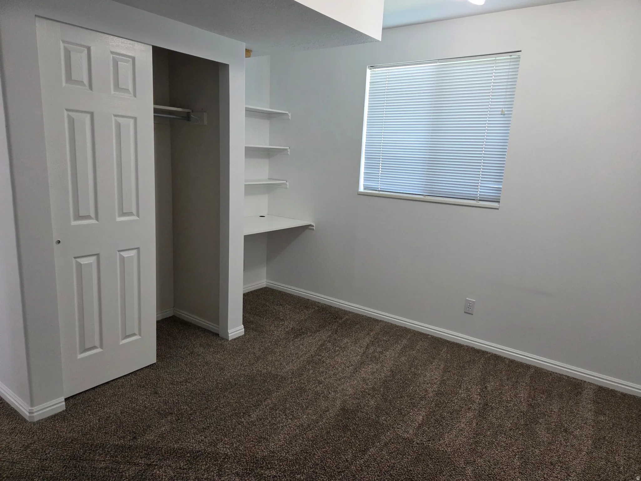 Carpeted room featuring a built-in desk with shelving, a window with blinds, and a closet with a bi-fold door