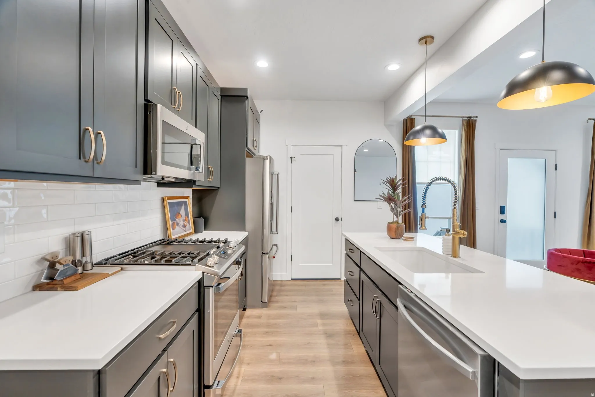 Modern kitchen featuring dark grey cabinetry with gold-toned hardware, stainless steel appliances, a white subway tile backsplash, and light wood-finish flooring