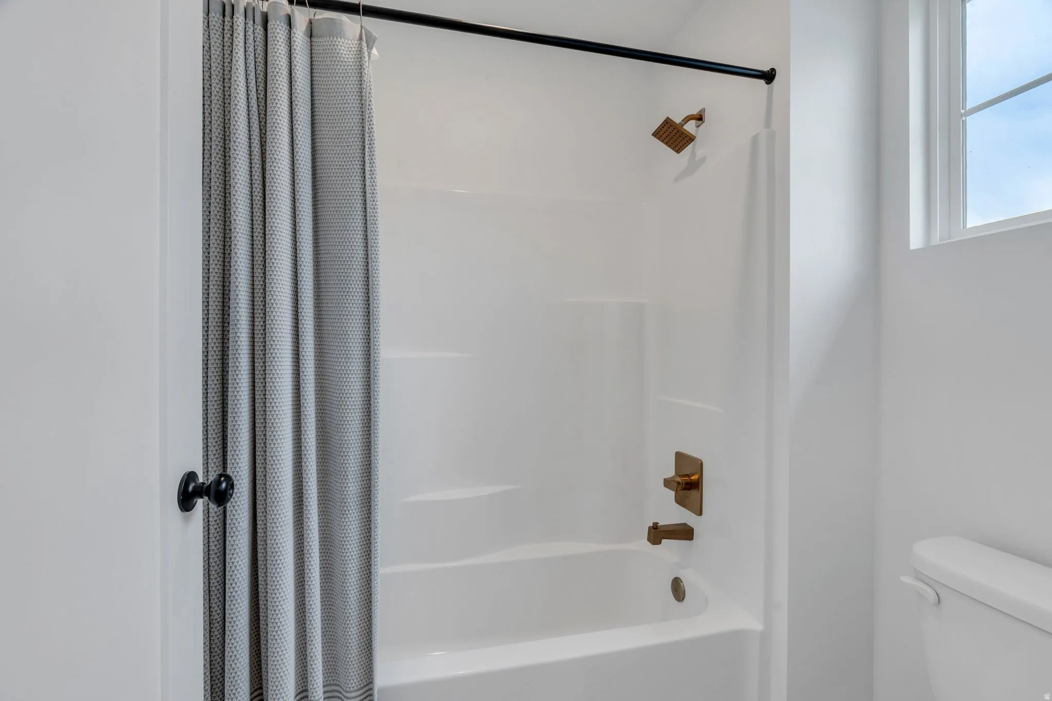 White shower/tub combination with brass fixtures, a black shower curtain rod, and a privacy window