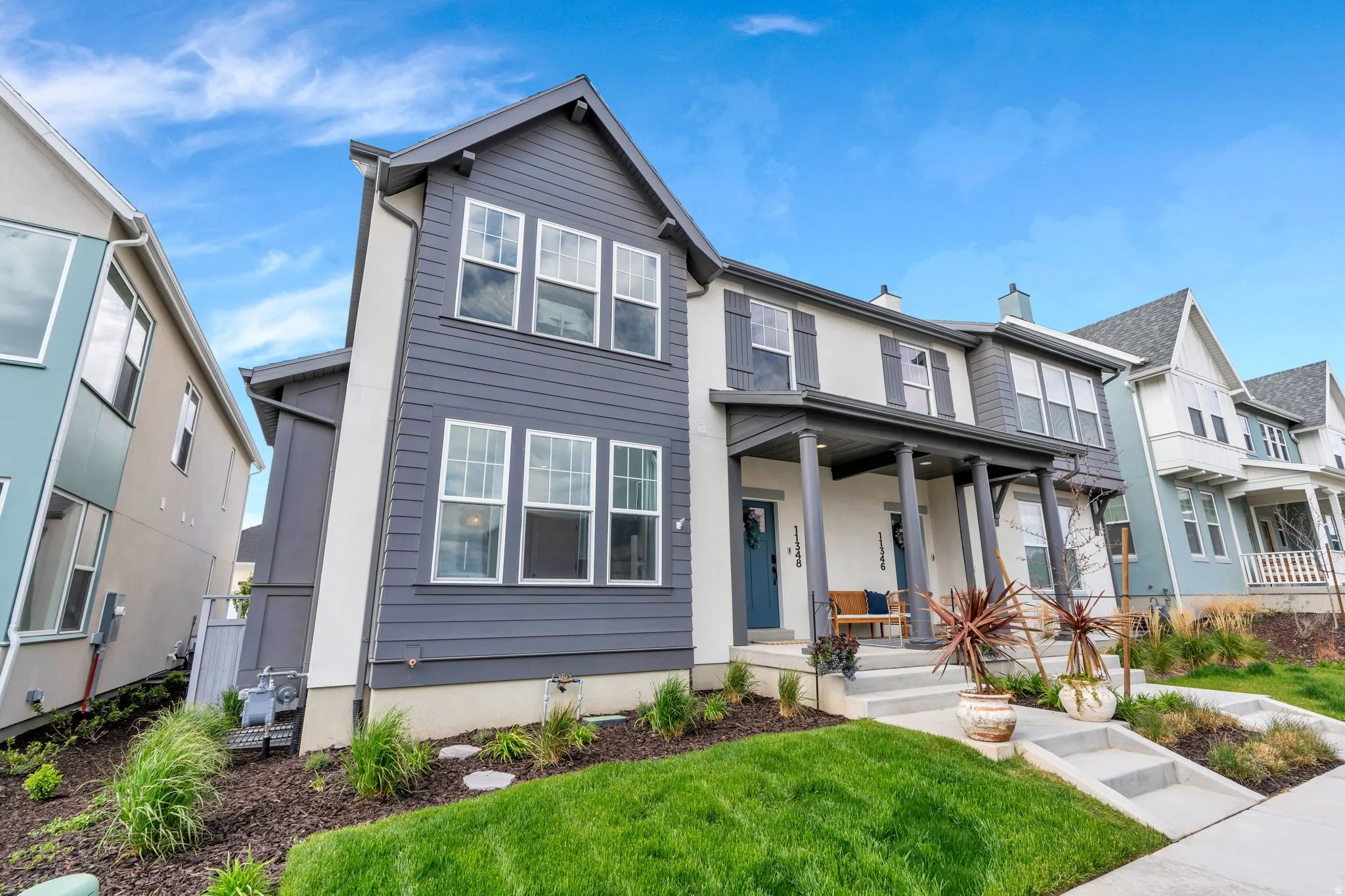 Modern two-story exterior featuring a prominent front porch with columns, a combination of horizontal siding and stucco finishes, multiple windows with dark shutters, and a landscaped front yard with green lawn and planted beds