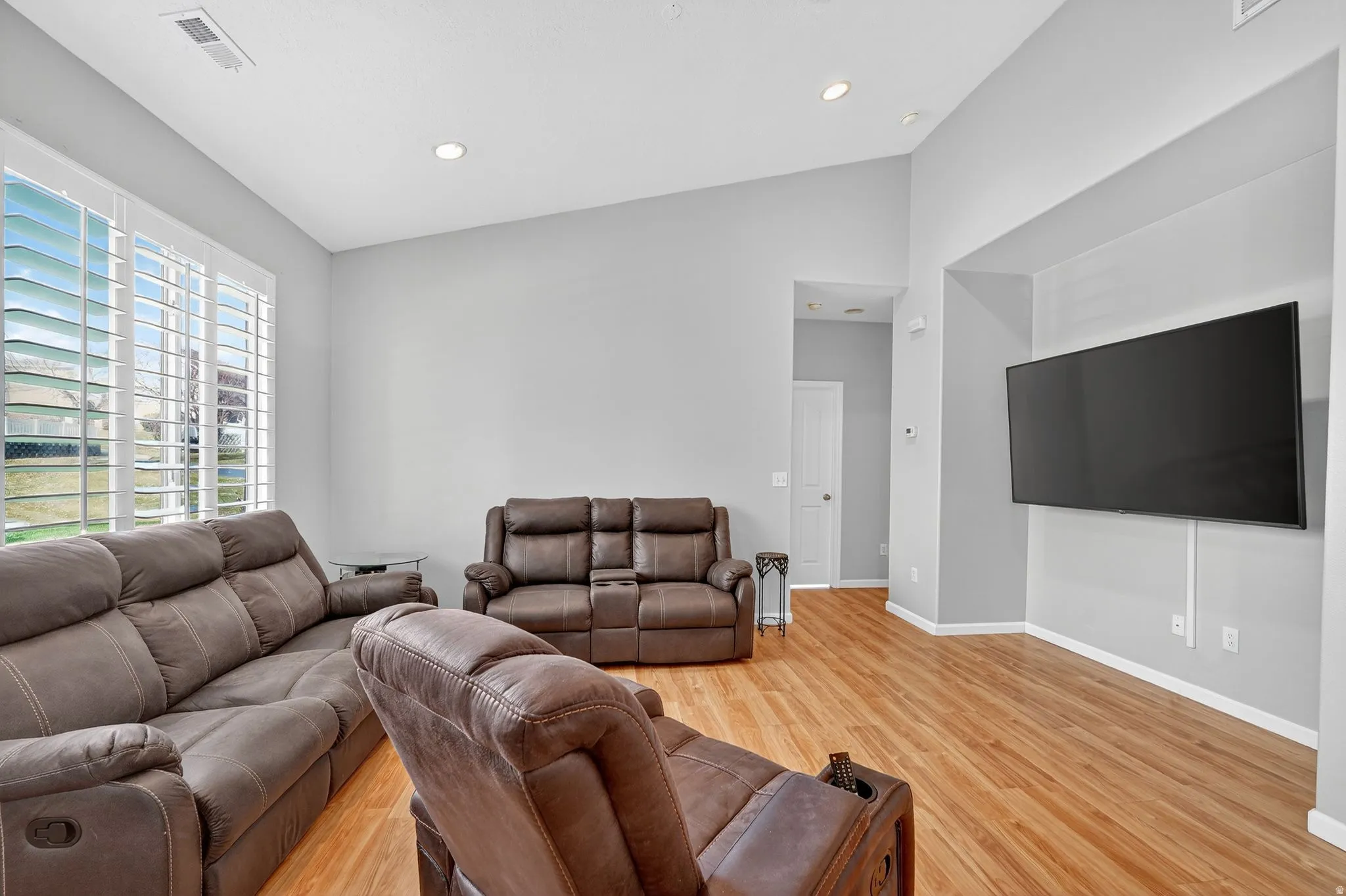 Spacious living area featuring wood-finish flooring, vaulted ceilings, recessed lighting, and a large window with plantation shutters