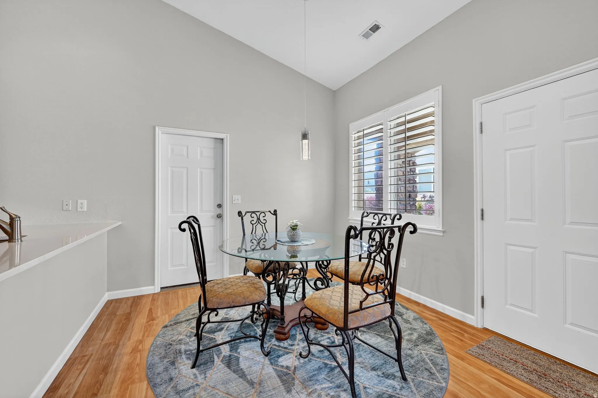 Vaulted ceiling dining area featuring light wood-finish flooring, white plantation shutters, a modern pendant light fixture, and light gray wall paint