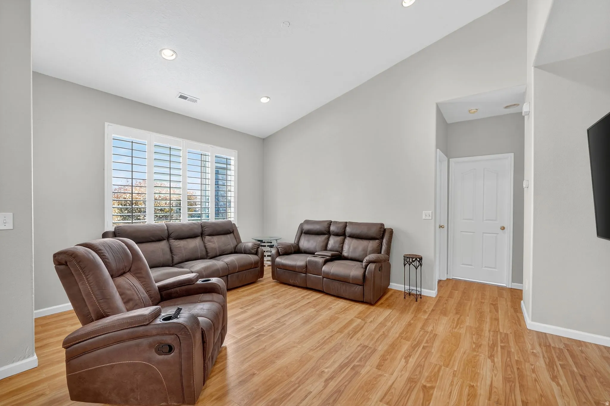 Spacious living area featuring wood-finish flooring, a vaulted ceiling with recessed lighting, and large windows with plantation shutters