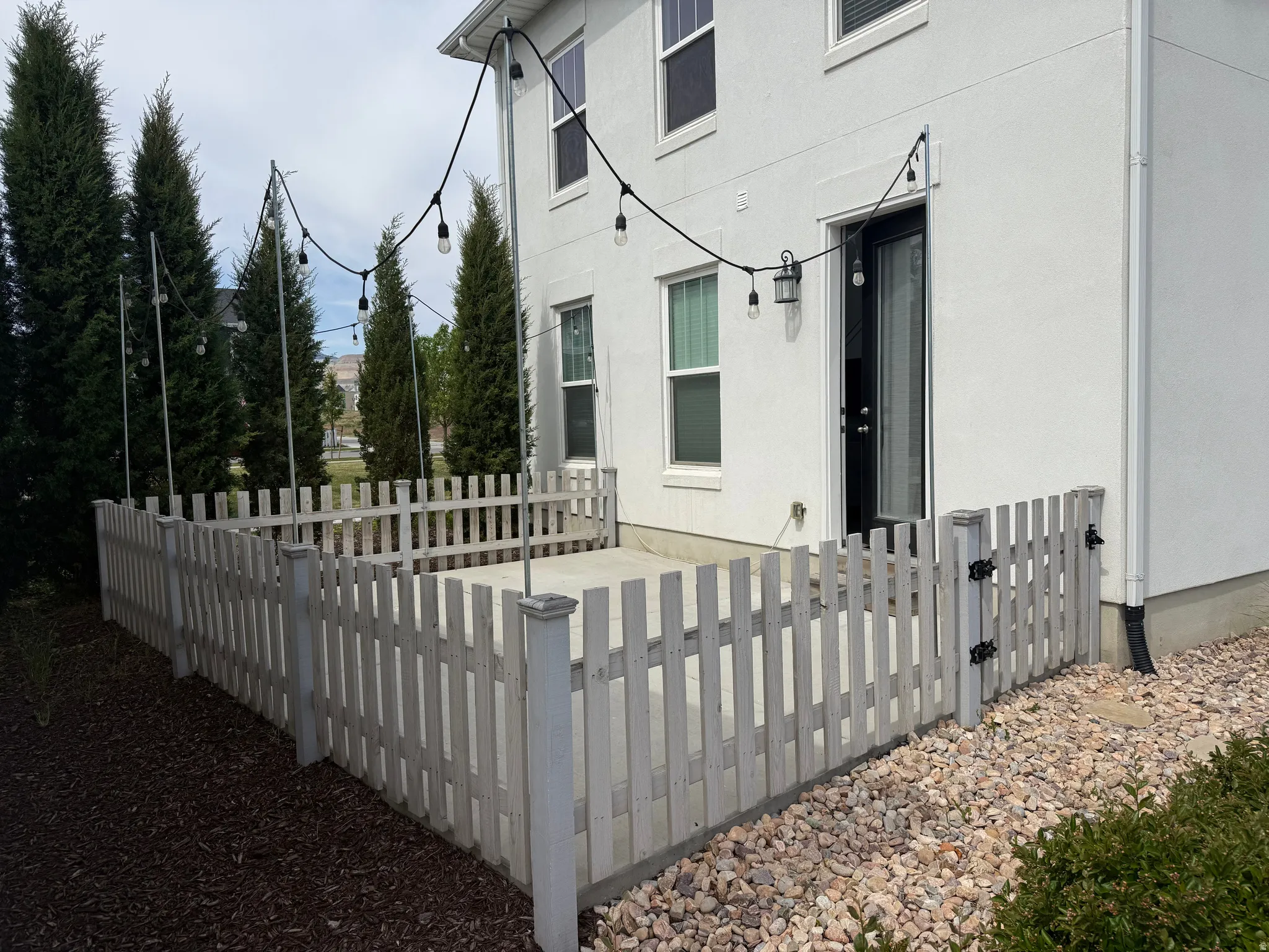 Enclosed patio featuring a light-toned picket fence, strung accent lighting, and a private entrance