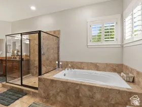 Spacious bathroom featuring a built-in jetted tub with stone surround, a separate glass-enclosed shower, dual vanity with wood cabinetry, tile flooring, and plantation shutters on multiple windows