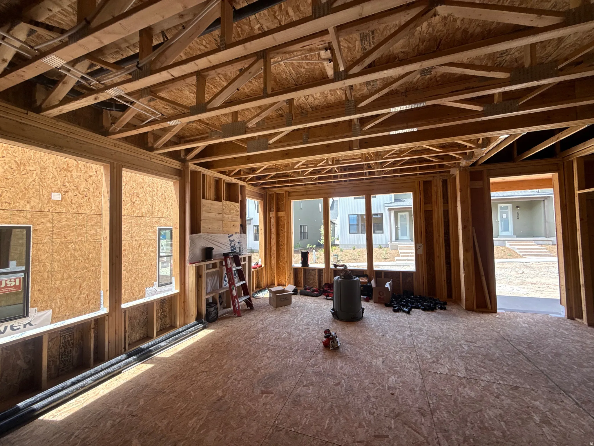 Framed interior space featuring exposed wood framing, ceiling trusses, large window openings, and oriented strand board subflooring