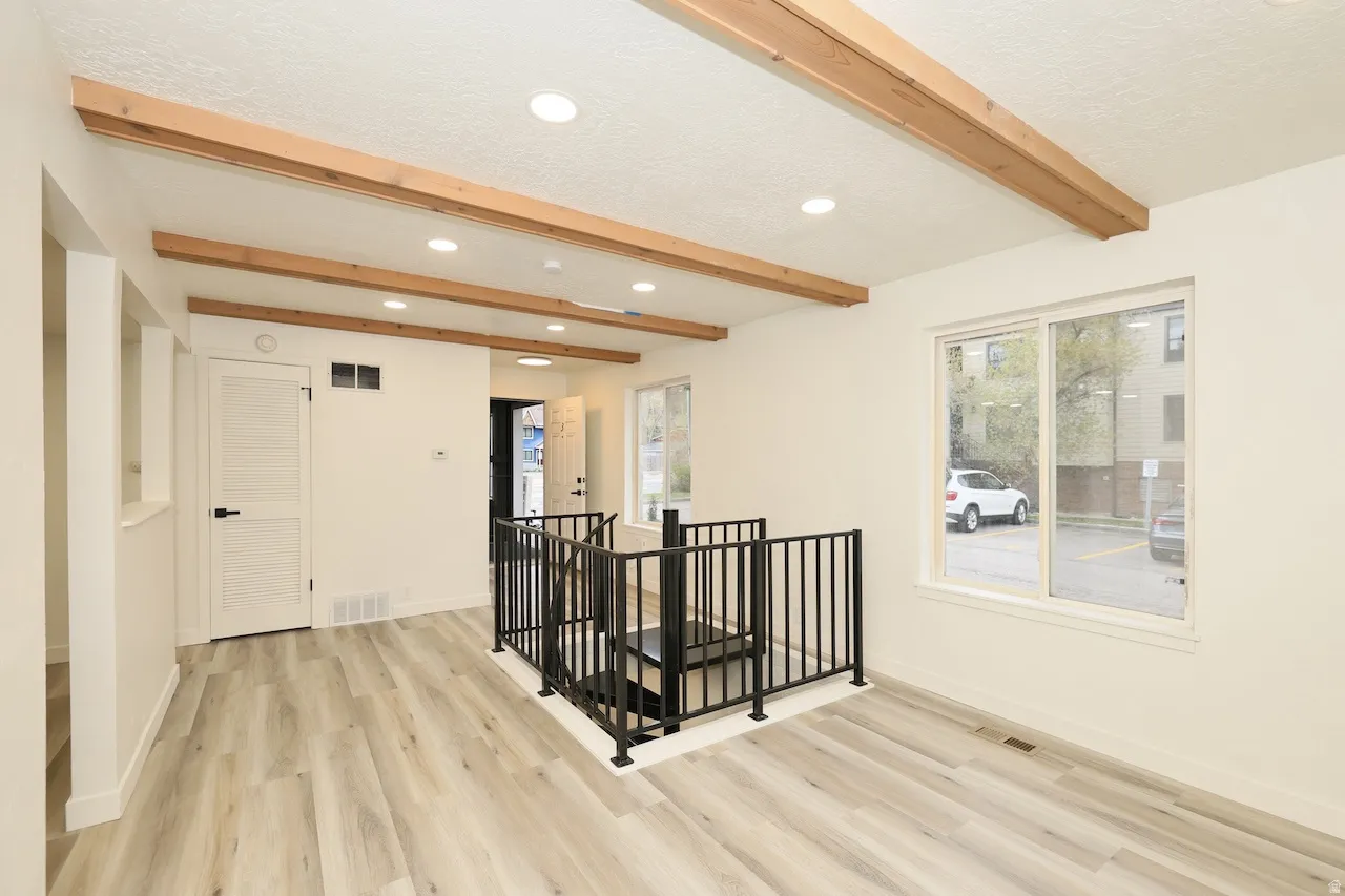 Open-concept living area featuring wood-finish flooring, exposed ceiling beams, recessed lighting, and a contemporary black metal stair railing