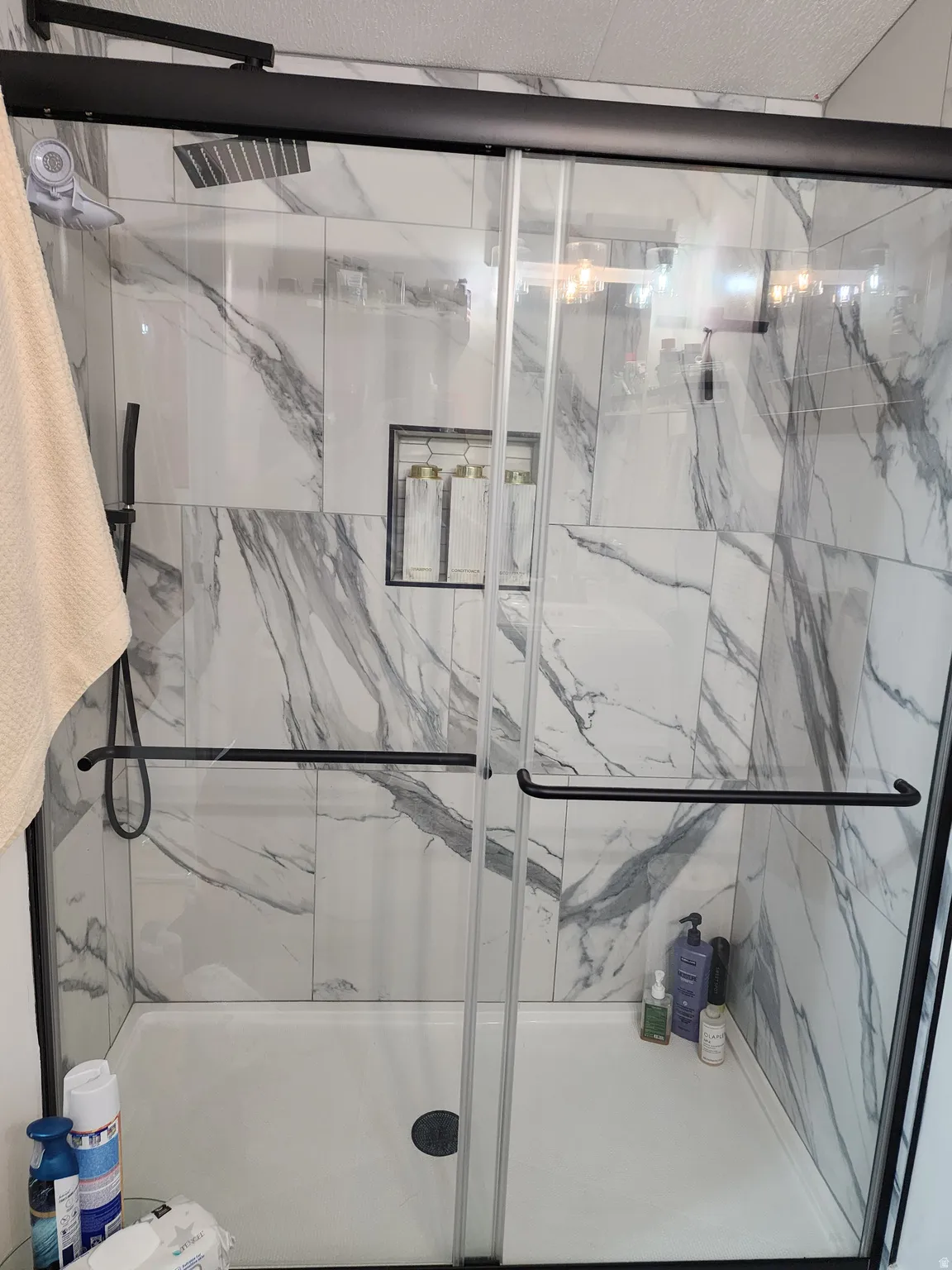 Walk-in shower featuring a sliding glass door with matte black hardware, large format marble-look tile, a built-in shower niche with mosaic tile, and a rain shower head