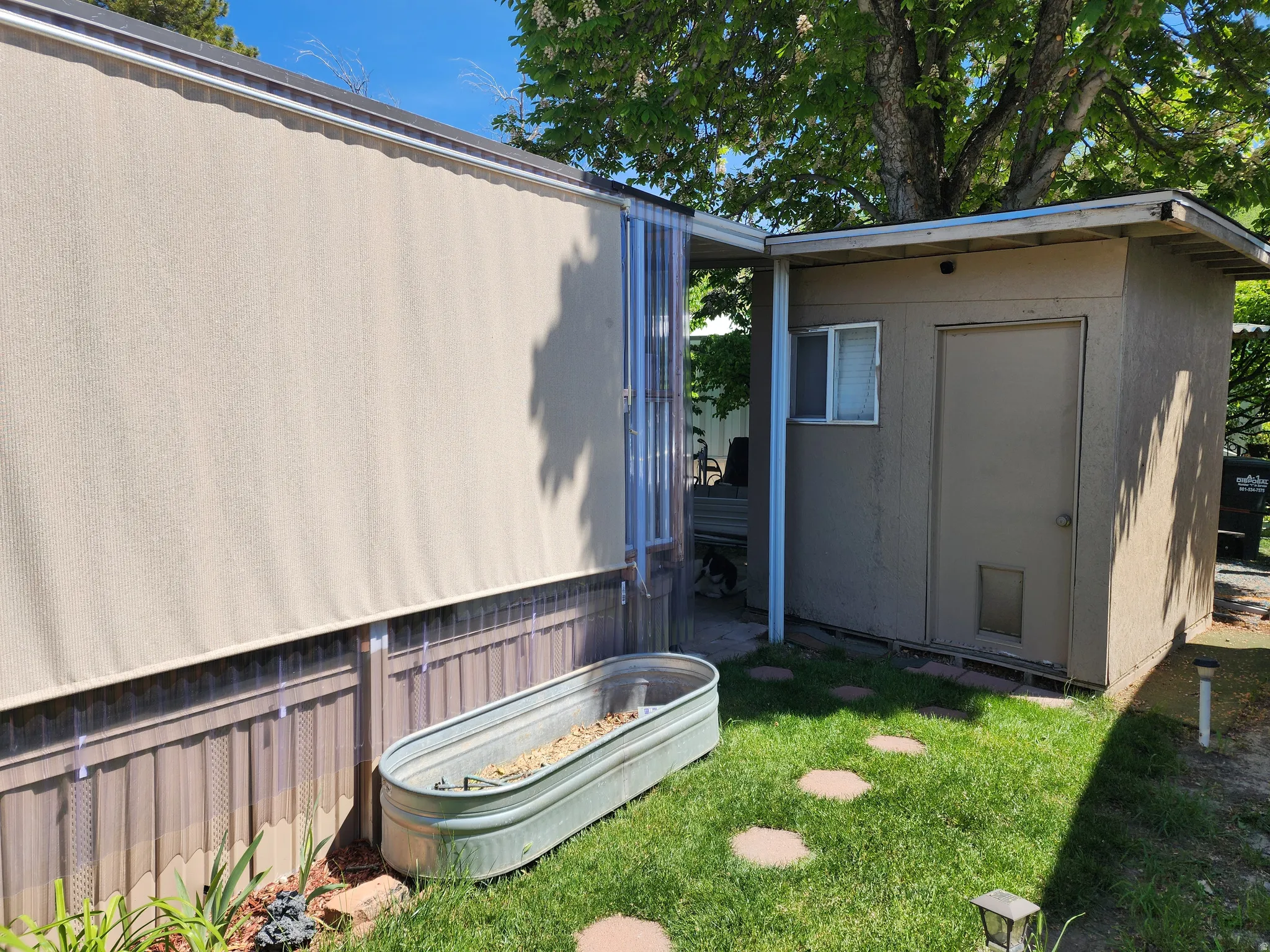 Exterior featuring a neutral-toned awning, corrugated perimeter skirting, a detached storage building with a window and door, and a grass area with stepping stones