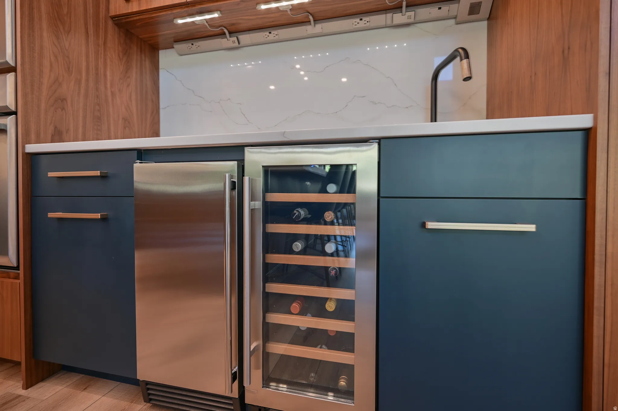 Integrated wet bar featuring a stainless steel beverage refrigerator, a glass-front wine cooler, a dark bronze faucet, and under-cabinet task lighting