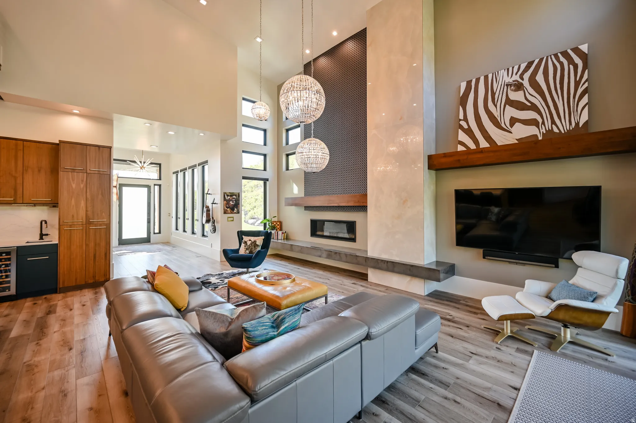 Double-height great room featuring a linear fireplace set within a textured accent wall, wood-finish flooring, a built-in wet bar with a beverage cooler, and expansive windows