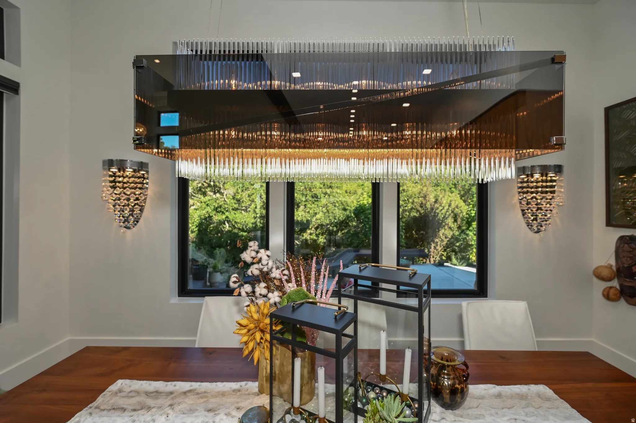 Contemporary dining area featuring a custom rectangular chandelier, two complementary wall sconces, expansive black-framed windows, and light-toned walls