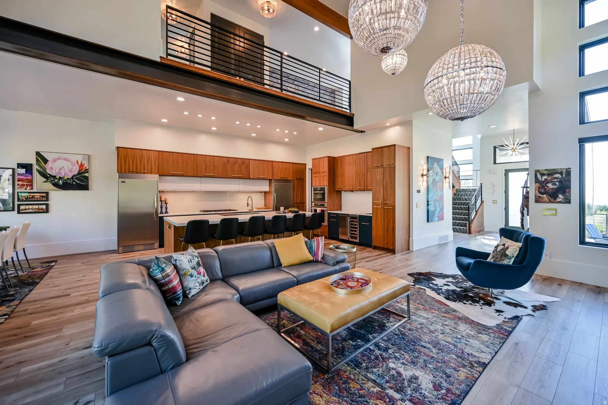Spacious open-concept living area featuring high ceilings, wood-finish flooring, a contemporary kitchen with stainless steel appliances, a kitchen island with seating, and a second-story interior balcony with an industrial-style railing