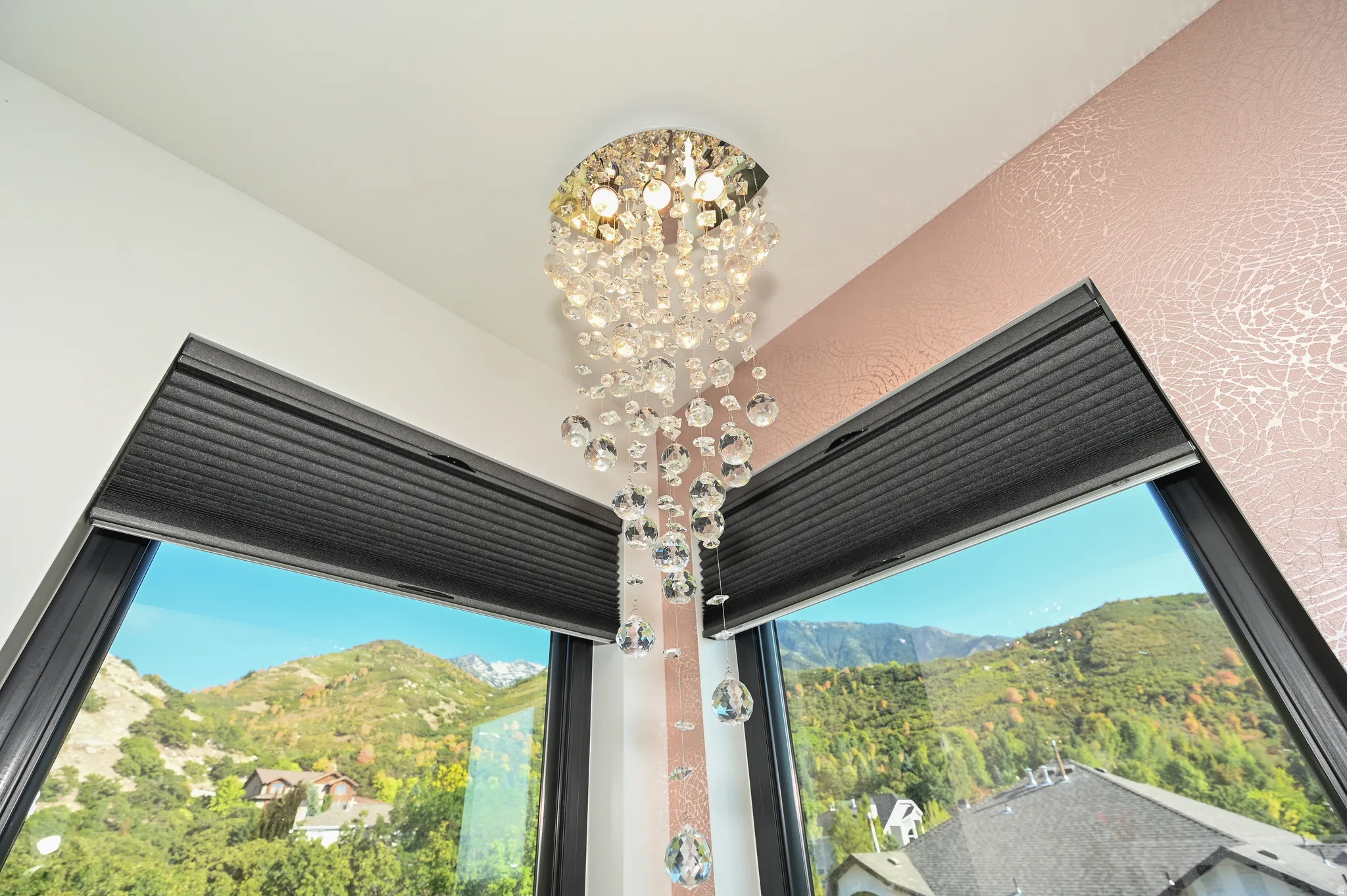 Corner window ensemble featuring a crystal chandelier, black framed windows, dark pleated blinds, and a distinctive textured wall finish
