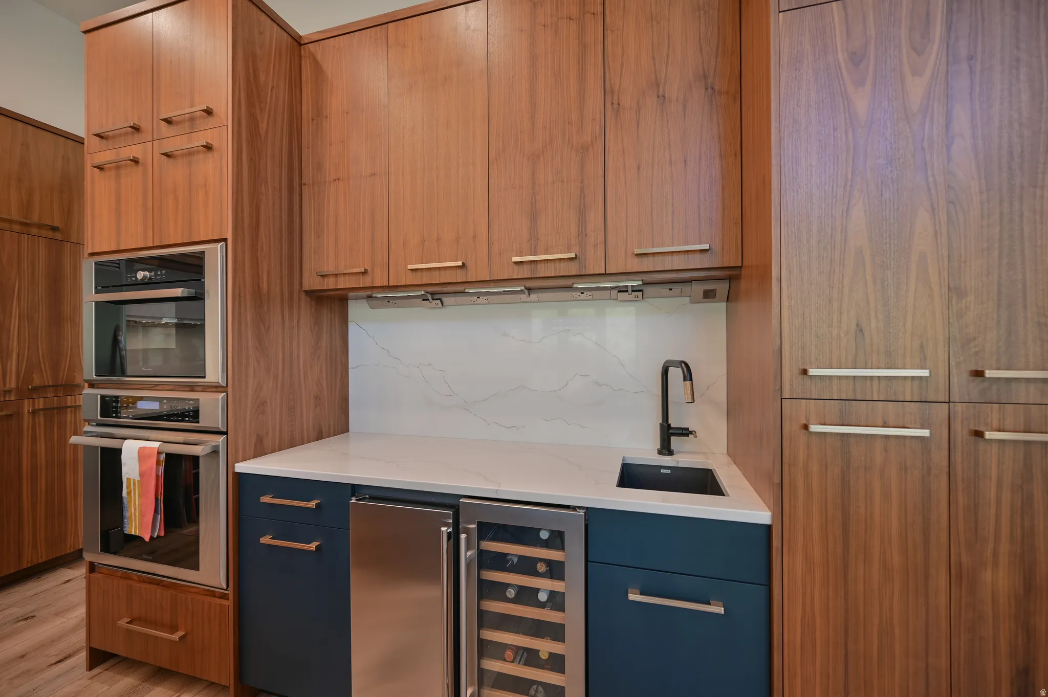 Built-in stainless steel double oven, wood-finish cabinetry, white countertop, integrated beverage refrigerator, and a matte black faucet with a coordinating sink