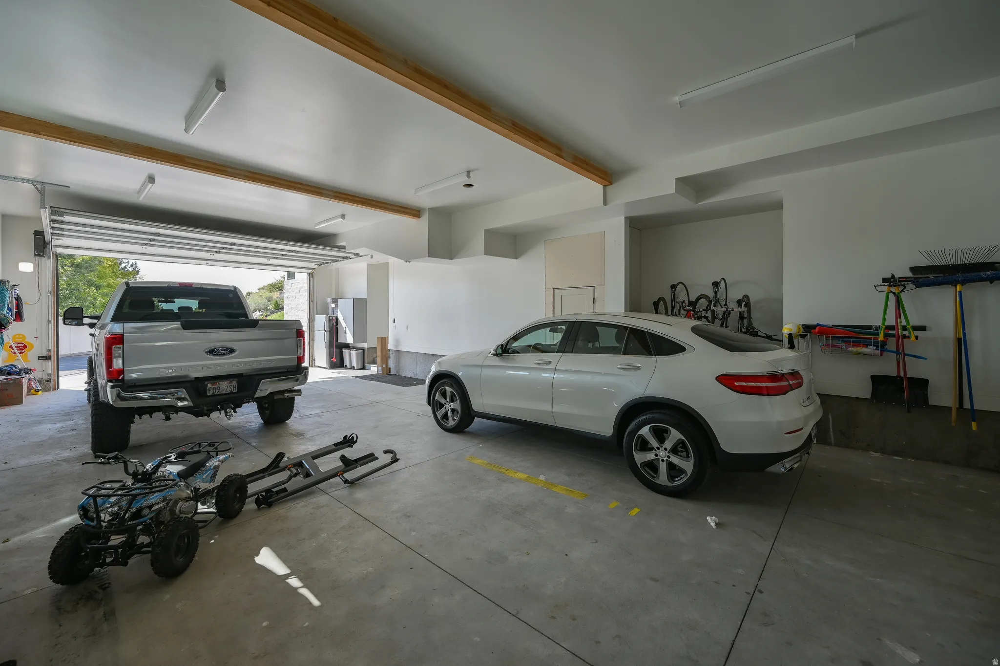 Spacious two-car garage featuring a concrete floor, white walls, exposed wood ceiling beams, and fluorescent strip lighting