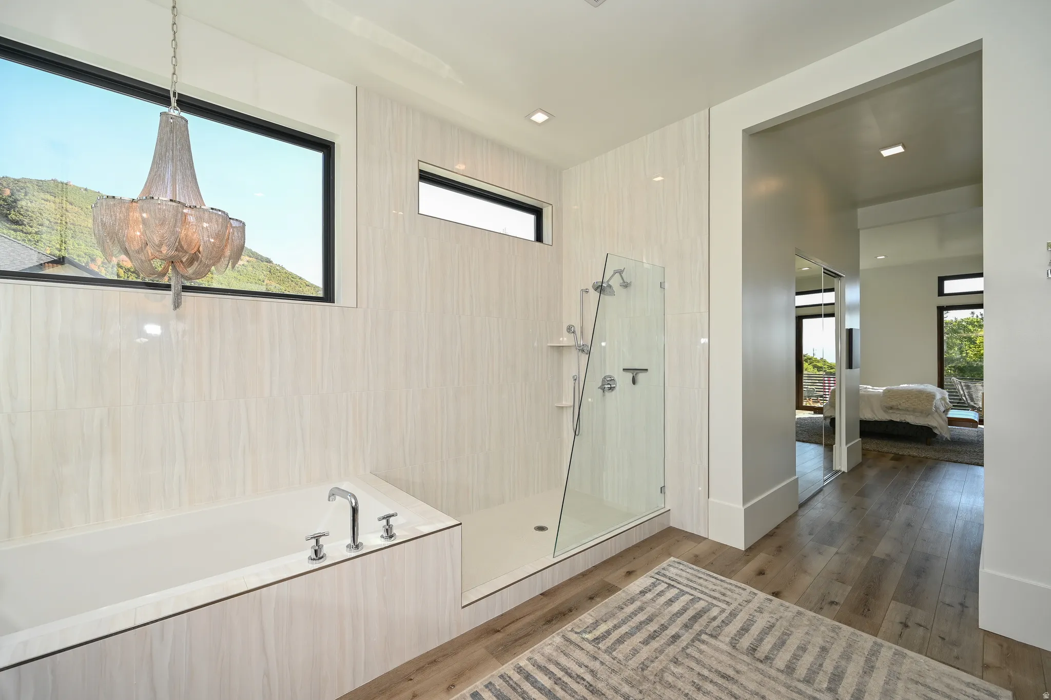 Spacious bathroom featuring a deep soaking tub with deck-mounted fixtures, a walk-in shower with glass enclosure, wood-finish flooring, and a decorative chandelier