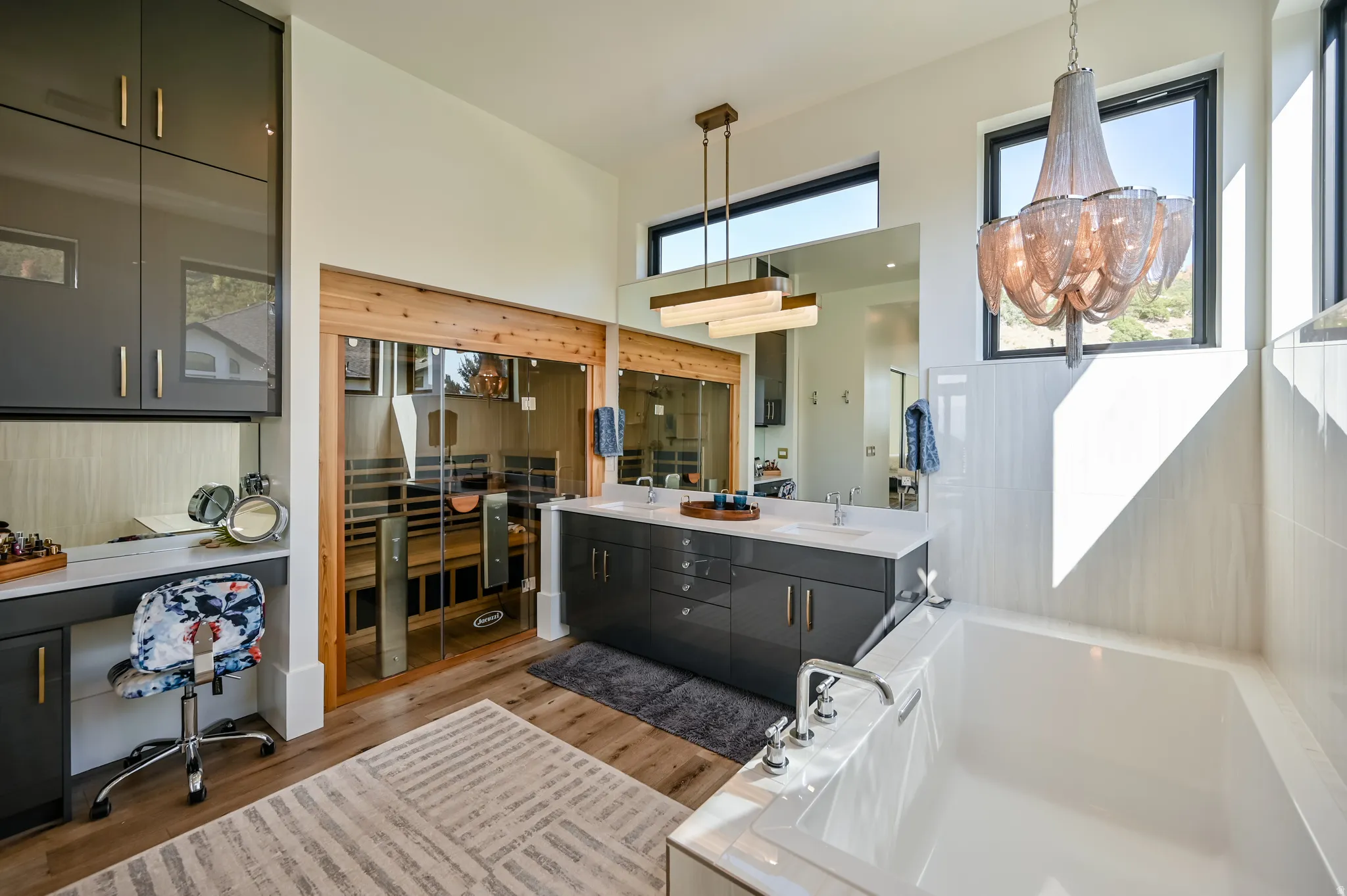 Spacious bathroom featuring a built-in sauna with wood-finish interior, a double vanity with undermount sinks, a built-in dressing table with upper cabinetry, a freestanding soaking tub, and wood-finish flooring