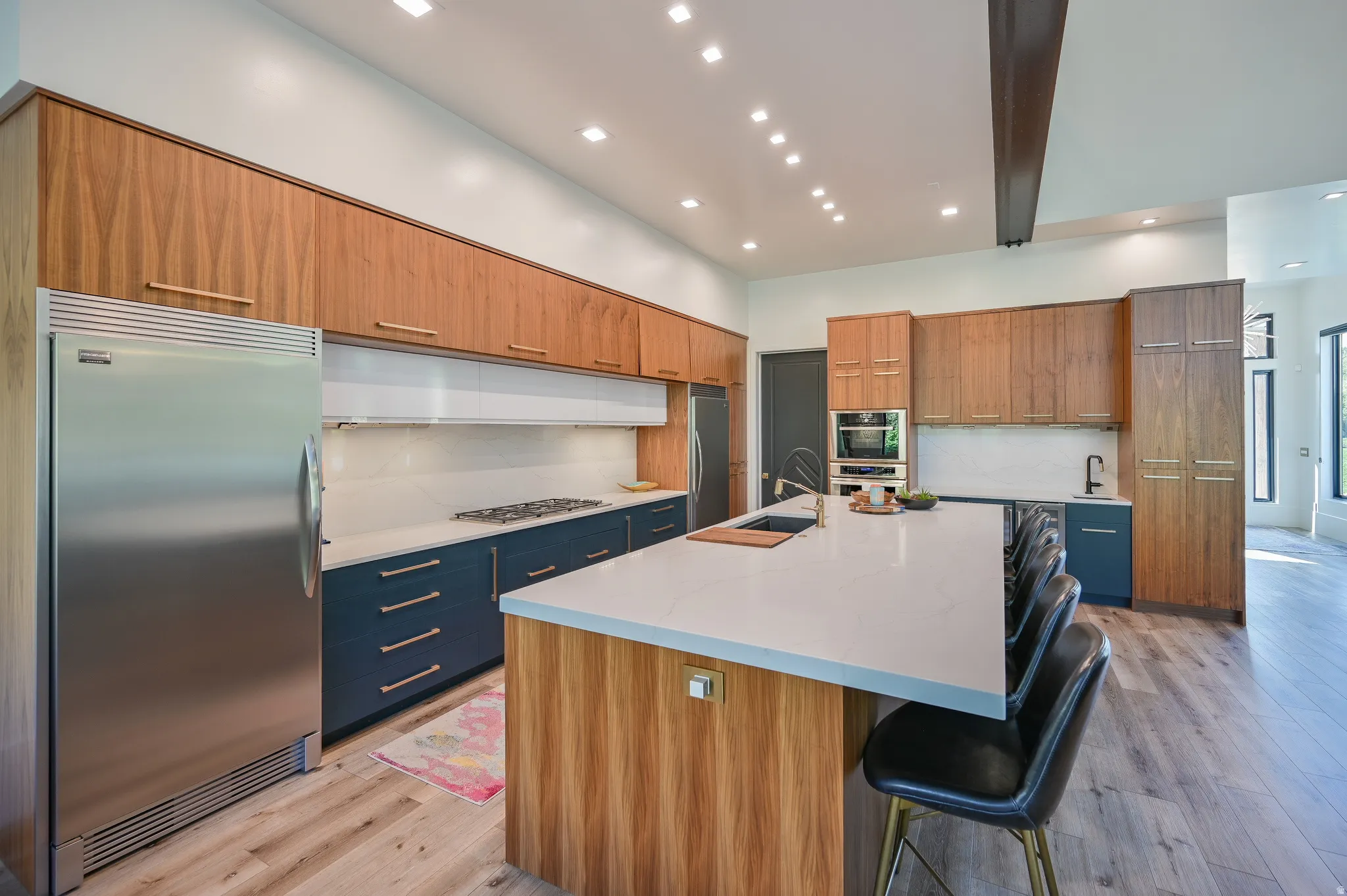 Modern kitchen featuring wood-finish upper cabinetry, lower cabinetry in a deep blue finish, stainless steel appliances, a center island with a light-toned countertop, and recessed lighting